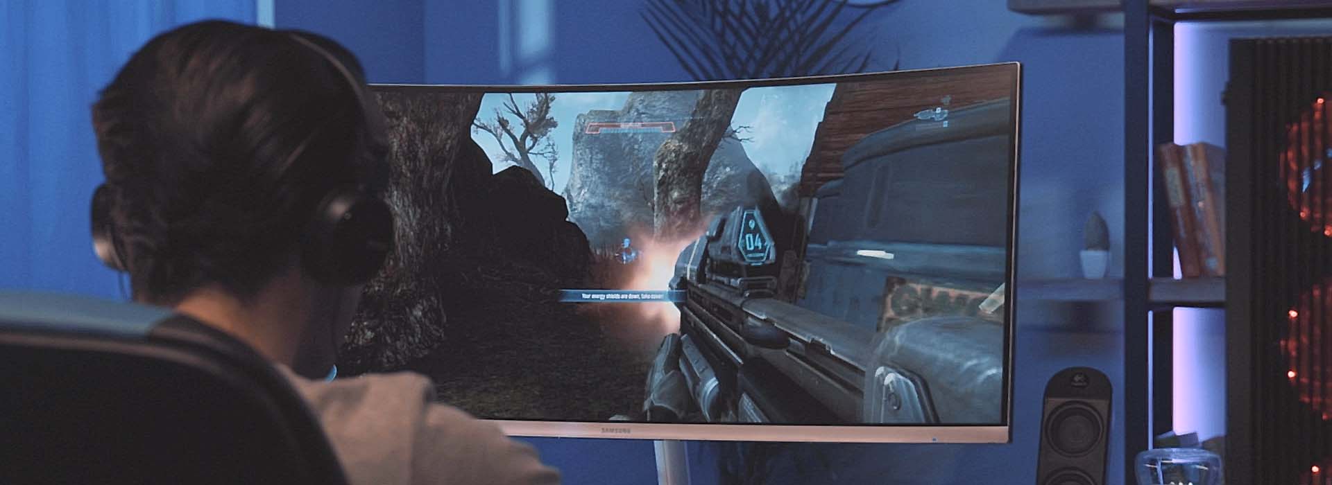 A man is seated in front of a Samsung gaming monitor playing a shooter game.